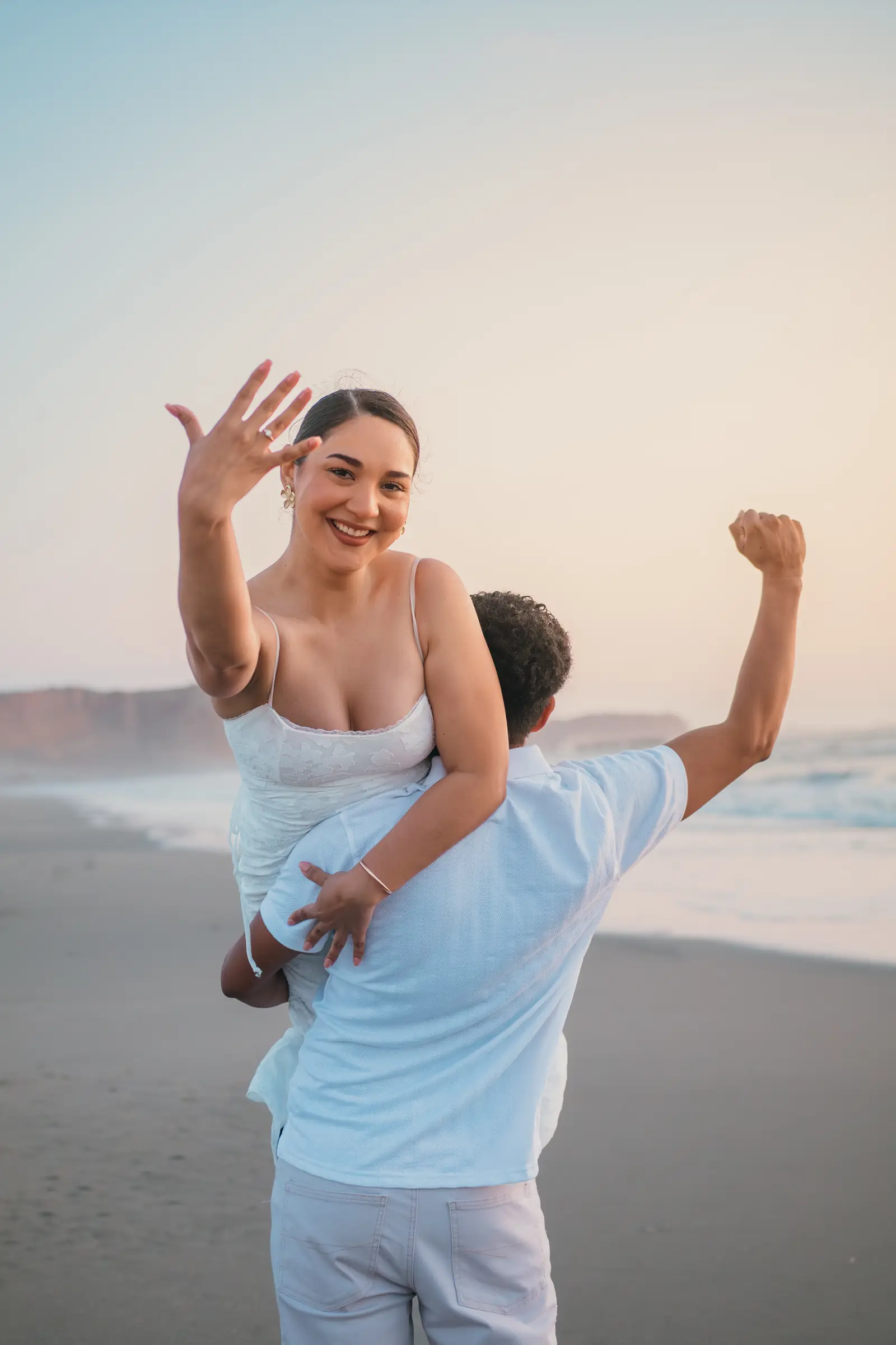 Mujer sonriendo con la mano en alto mostrando su anillo mientras su pareja la carga en la playa al atardecer